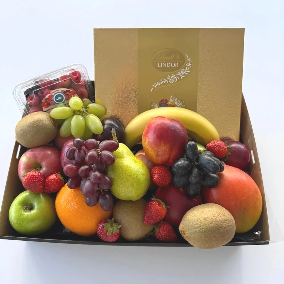 A box filled with assorted fresh fruits including strawberries, grapes, kiwis, apples, pears, bananas, and peaches, with a Lindt Lindor chocolate box in the background.