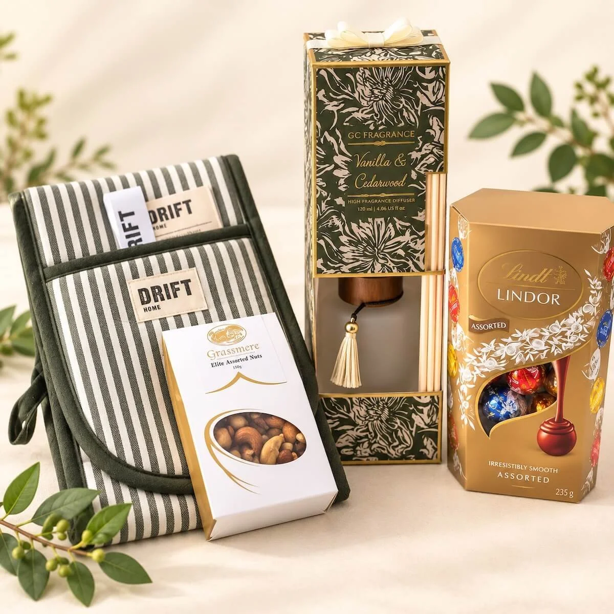 Housewarming gift accessories featuring a Drift striped oven mitt, assorted nuts, Lindt chocolates and Vanilla & Cedarwood fragrance diffuser.