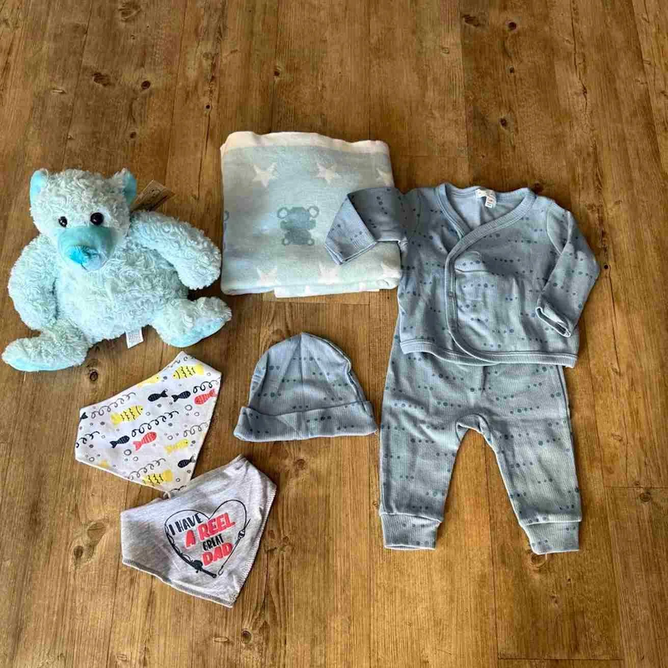 Baby clothing in athe jack hamper is a 3-piece blue set., pants, top and hat, a blue blanket & Teddy and two bibs.