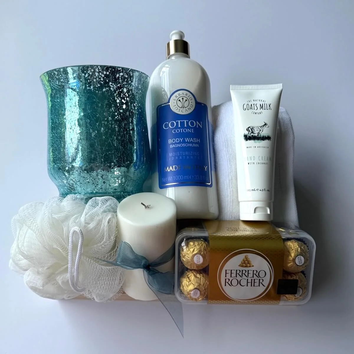 Luxury pamper gift hamper with Ferrero Rocher chocolates, goats milk hand cream, cotton body wash, candle, soft towel and blue glass candle holder.