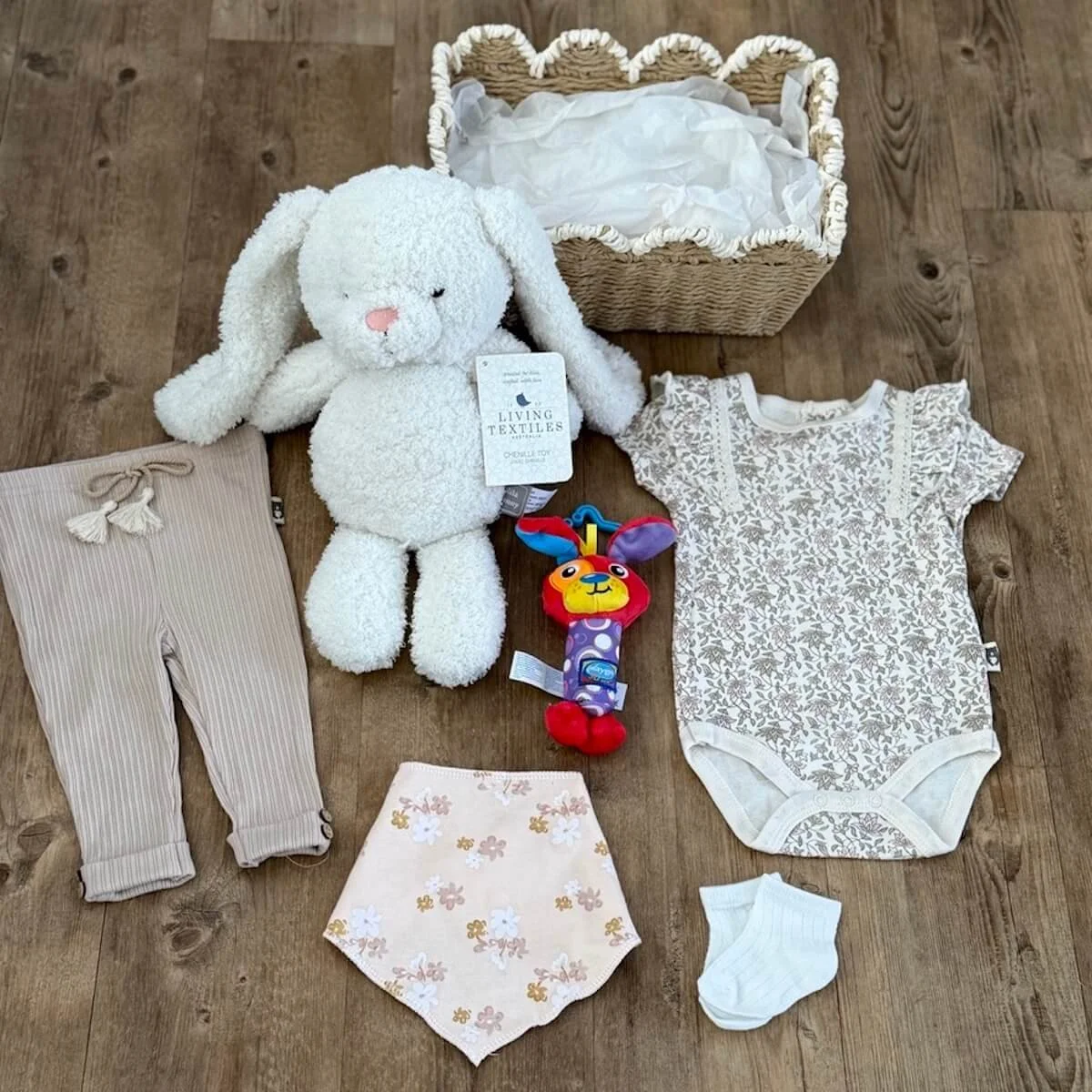 Baby girl gift set with floral onesie, cream pants, socks, bandana bib, bunny toy and pram rattle displayed outside woven basket