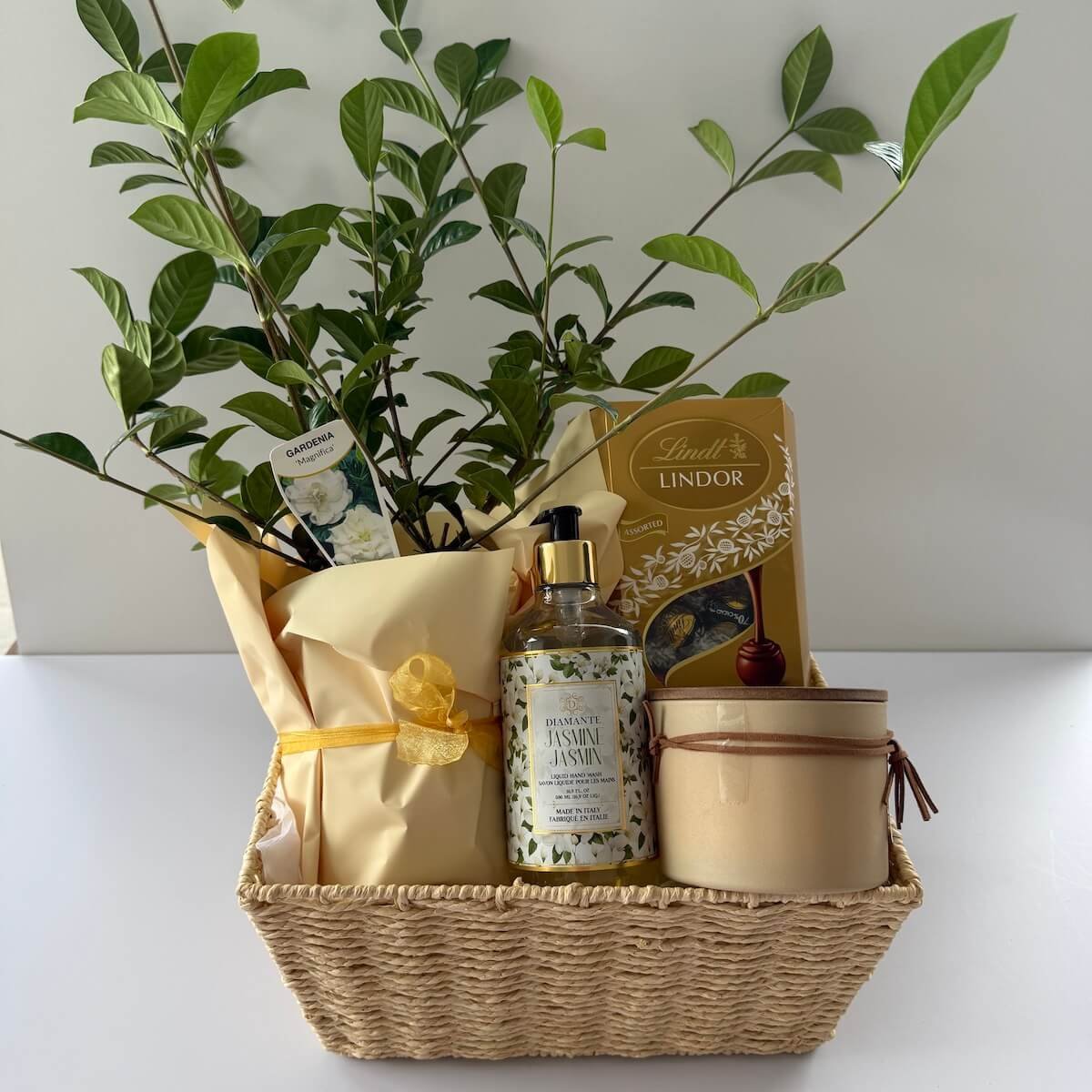 A gardenia plant that has scented flowers, a scented candle, hand wash and chocolates in a woven gift basket.