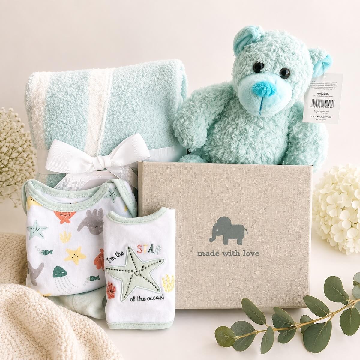The Little One Baby Hamper features a Blue baby Blanket, a Teddy bear, a photo album and a 3-piece 0-6 month clothing set.