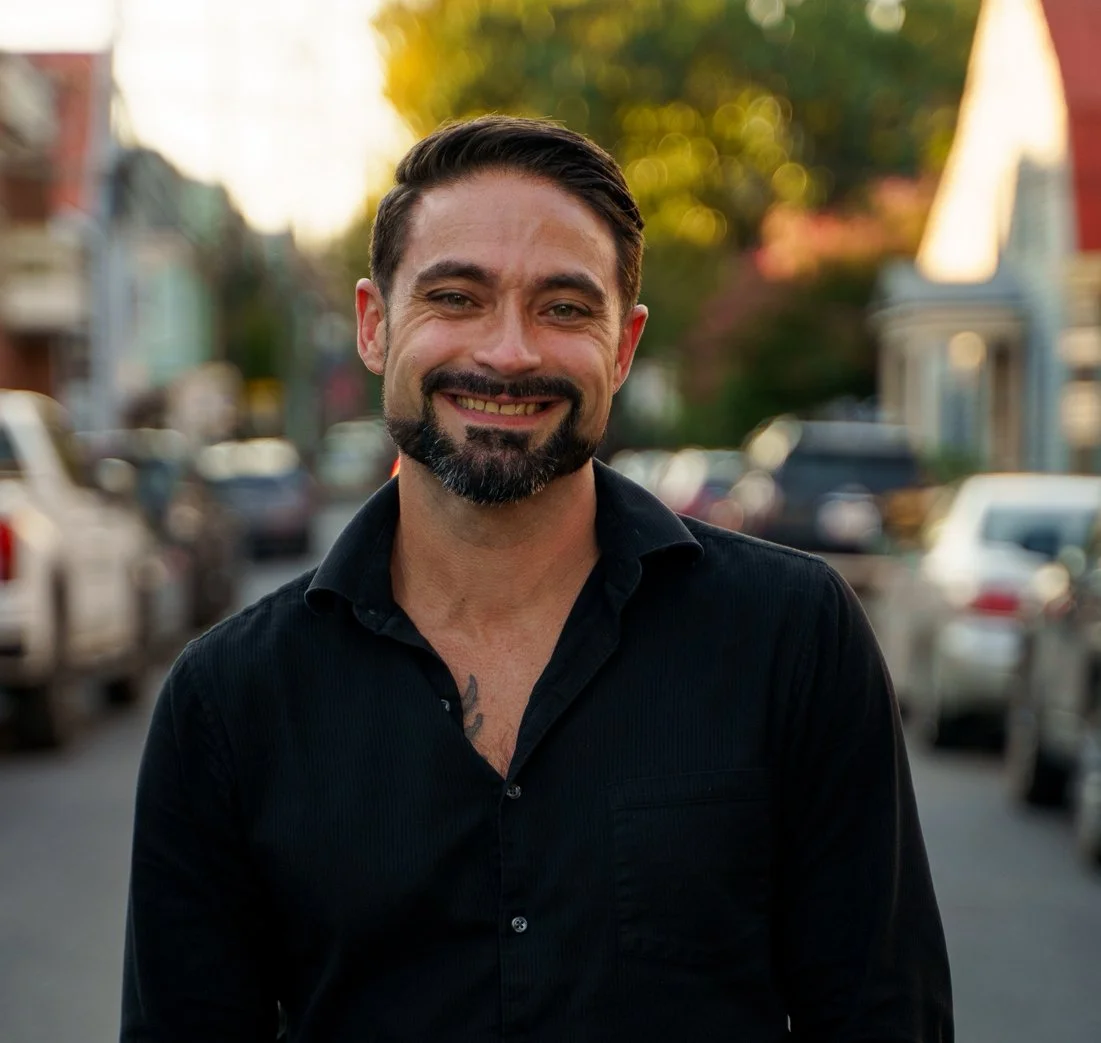 Smiling man with dark hair and beard wearing a black shirt, standing outdoors on a city street during golden hour with cars and buildings in the background.