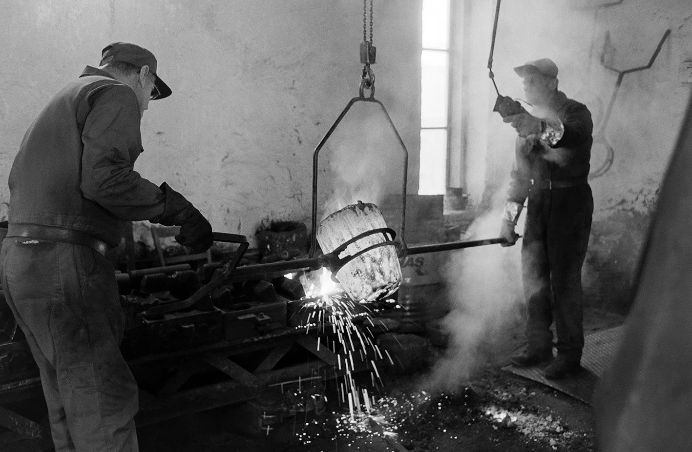 Two black and white photo of two men working with molten metal in a workshop. One man is pouring molten metal from a container into a mold, while the other is holding a tool and inspecting the process.