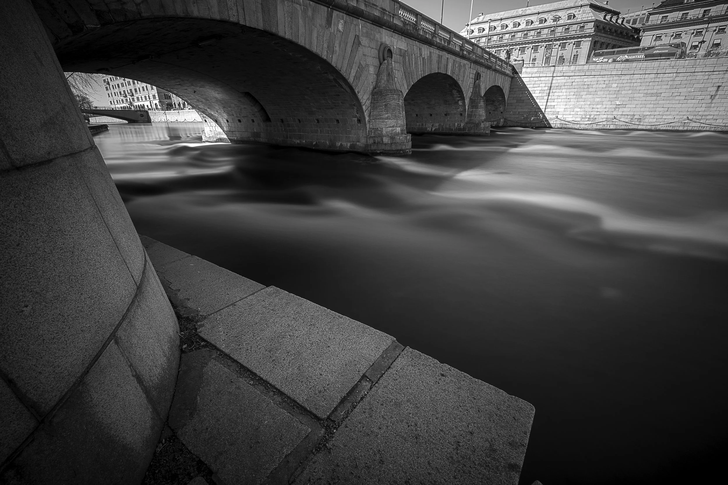 Black and white photograph of a stone bridge over a flowing river in an urban setting.