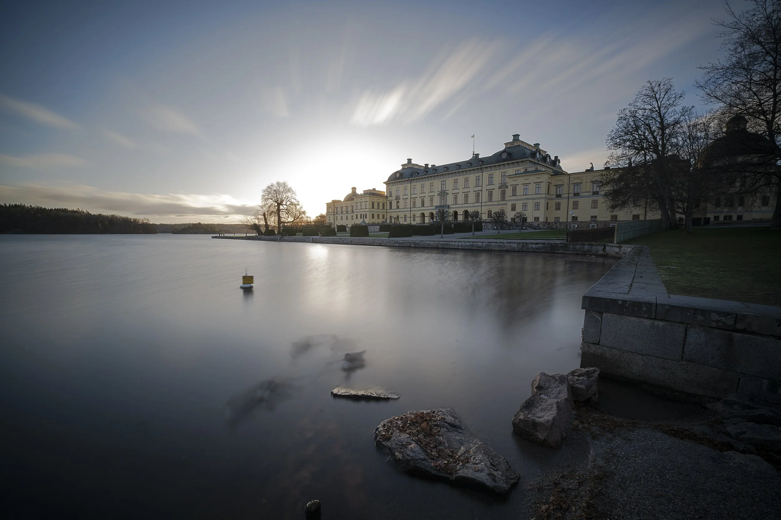 A large historic castle-like building with cream and yellow walls situated along a body of water, with leafless trees nearby and a cloudy sky backlit by the setting or rising sun.