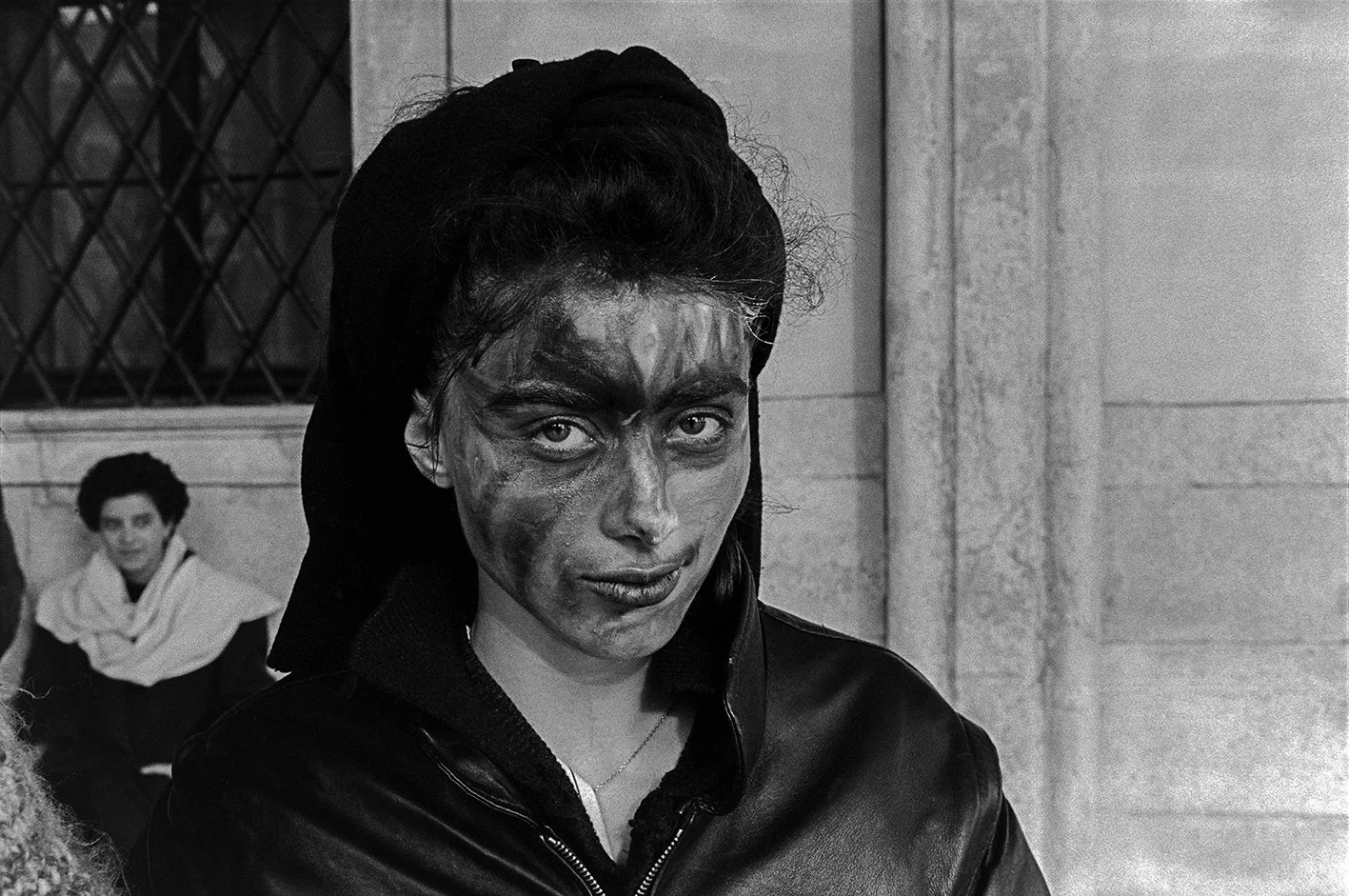 Black and white photo of a woman with smeared makeup or dirt on her face, wearing a jacket with the collar up, standing outdoors near a stone building, with a woman sitting in the background.
