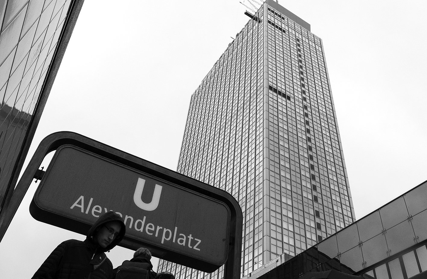 A black-and-white photo of Alexanderplatz subway station entrance with a large modern skyscraper in the background, and two people wearing hooded jackets near the station sign.
