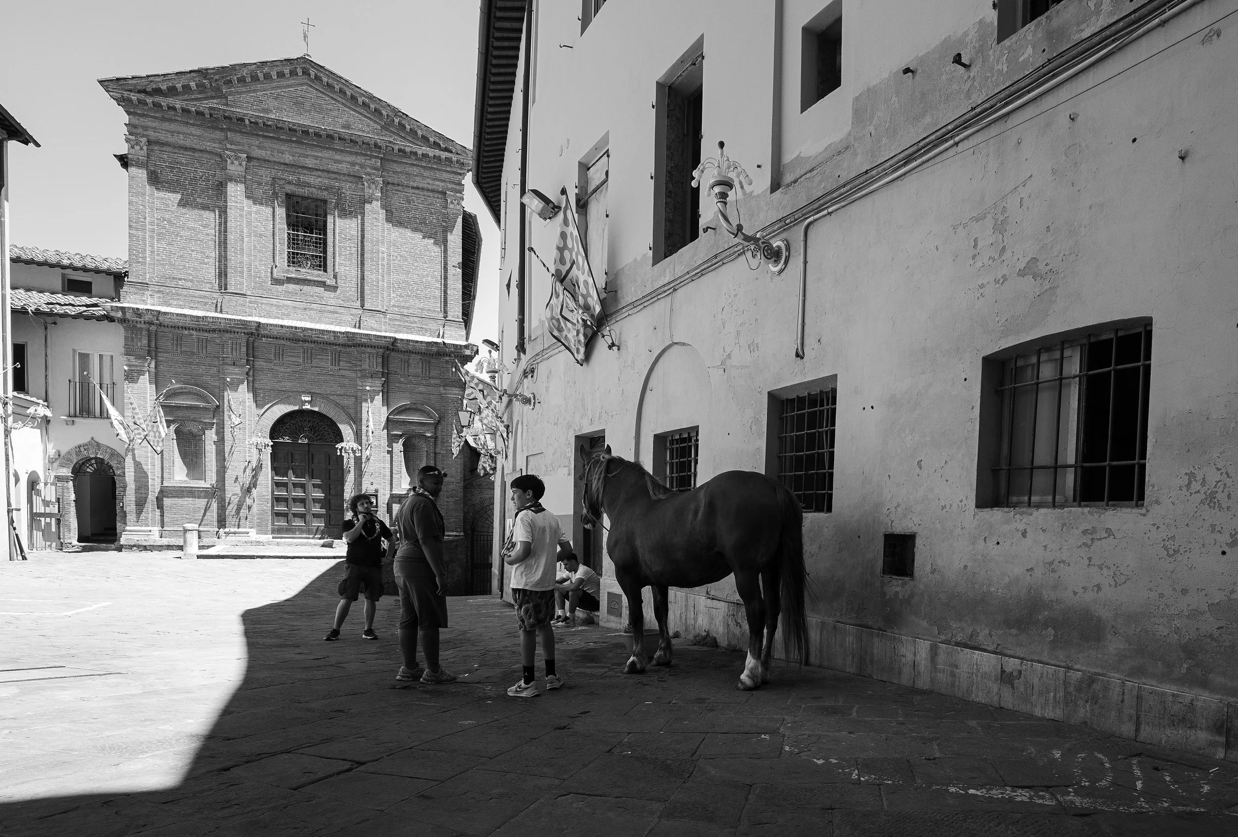 A black-and-white photo of a group of people and a horse standing in a historic town square with old brick and stucco buildings, banners, and a church in the background.