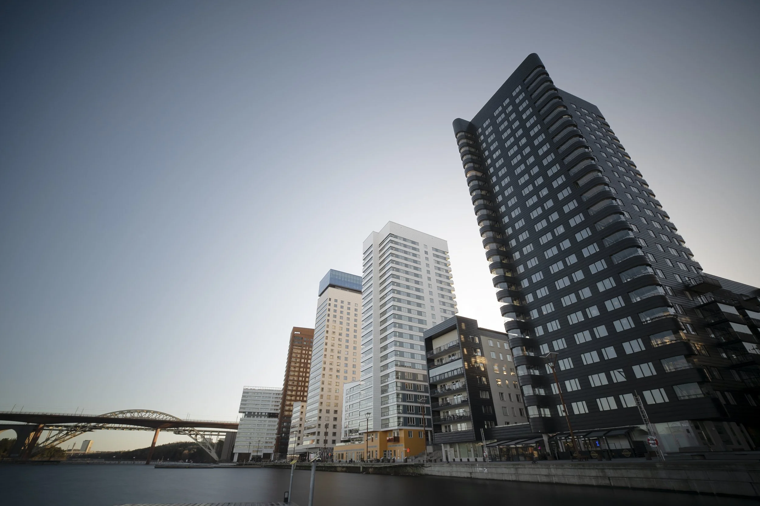 A cityscape of modern high-rise buildings along a river with a bridge in the background, under a clear sky.