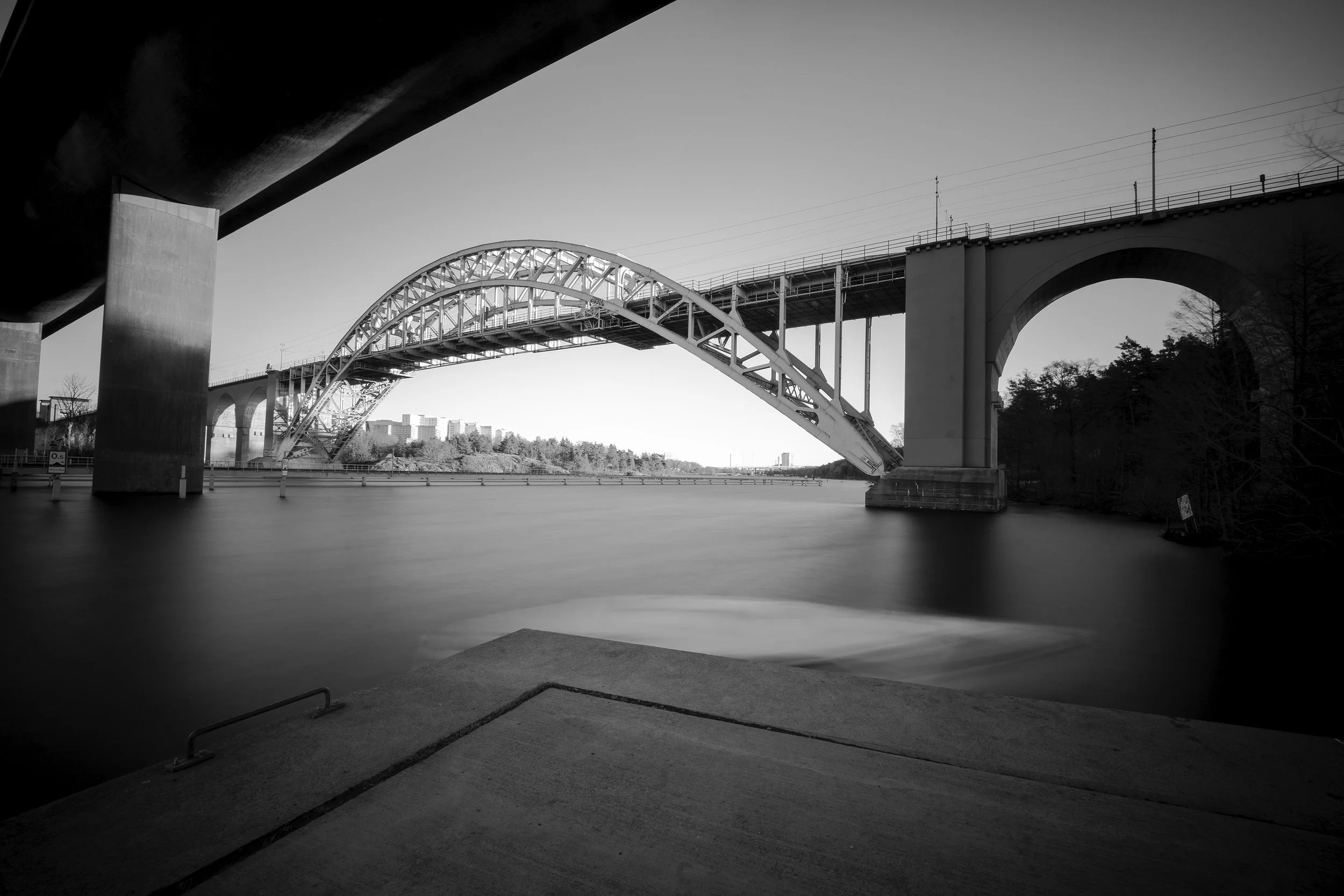Black and white photo of a bridge over a body of water, with an arch design and supporting columns, viewed from a concrete dock area.