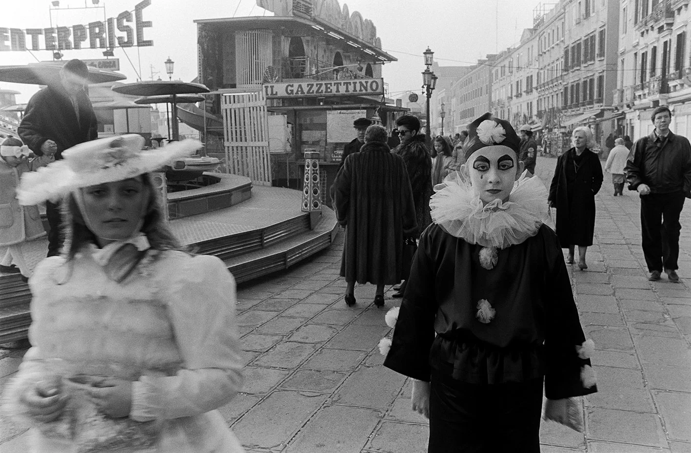 Two women walking on a busy street resembling Venice, Italy, one dressed as a clown with a ruffled collar and painted face, the other in period costume with a large hat, with a carnival atmosphere.