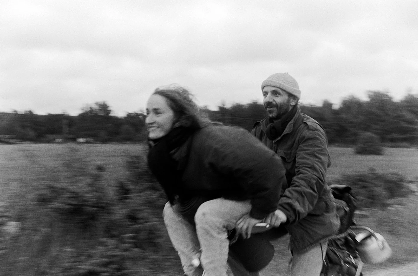 A man and a woman riding a motorcycle together outdoors on a cloudy day, with trees and open fields in the background.