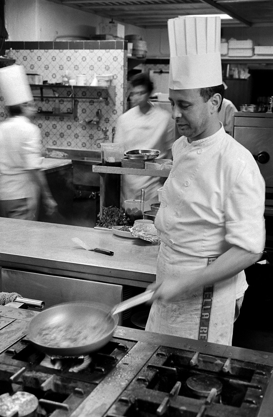 Chef cooking on stove in professional kitchen, with other kitchen staff in the background.