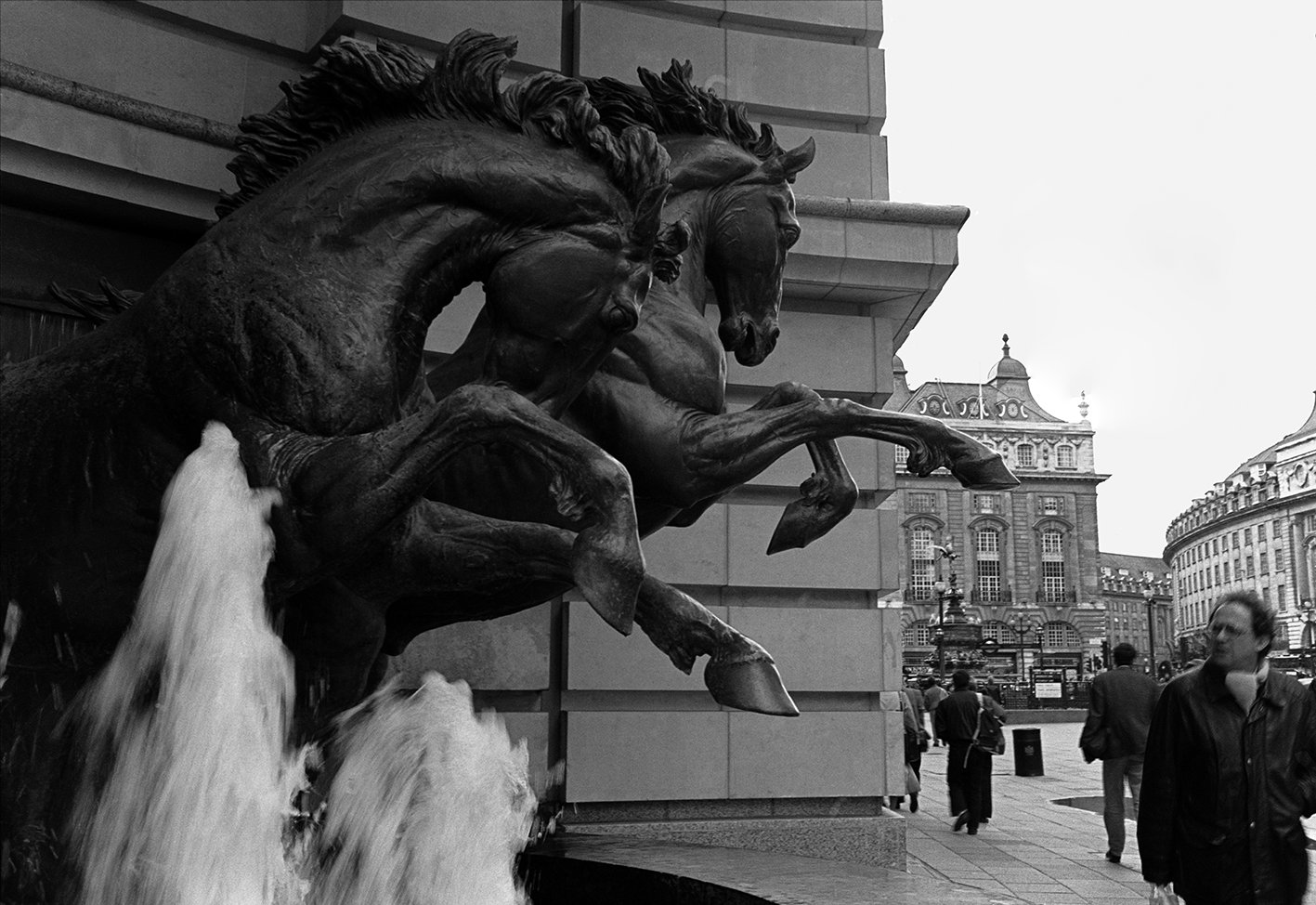 A black and white photo of a large bronze sculpture of three horses' heads and necks emerging from a building, with water flowing from their mouths into a basin below. People walk on the sidewalk nearby, and ornate buildings are visible in the background.