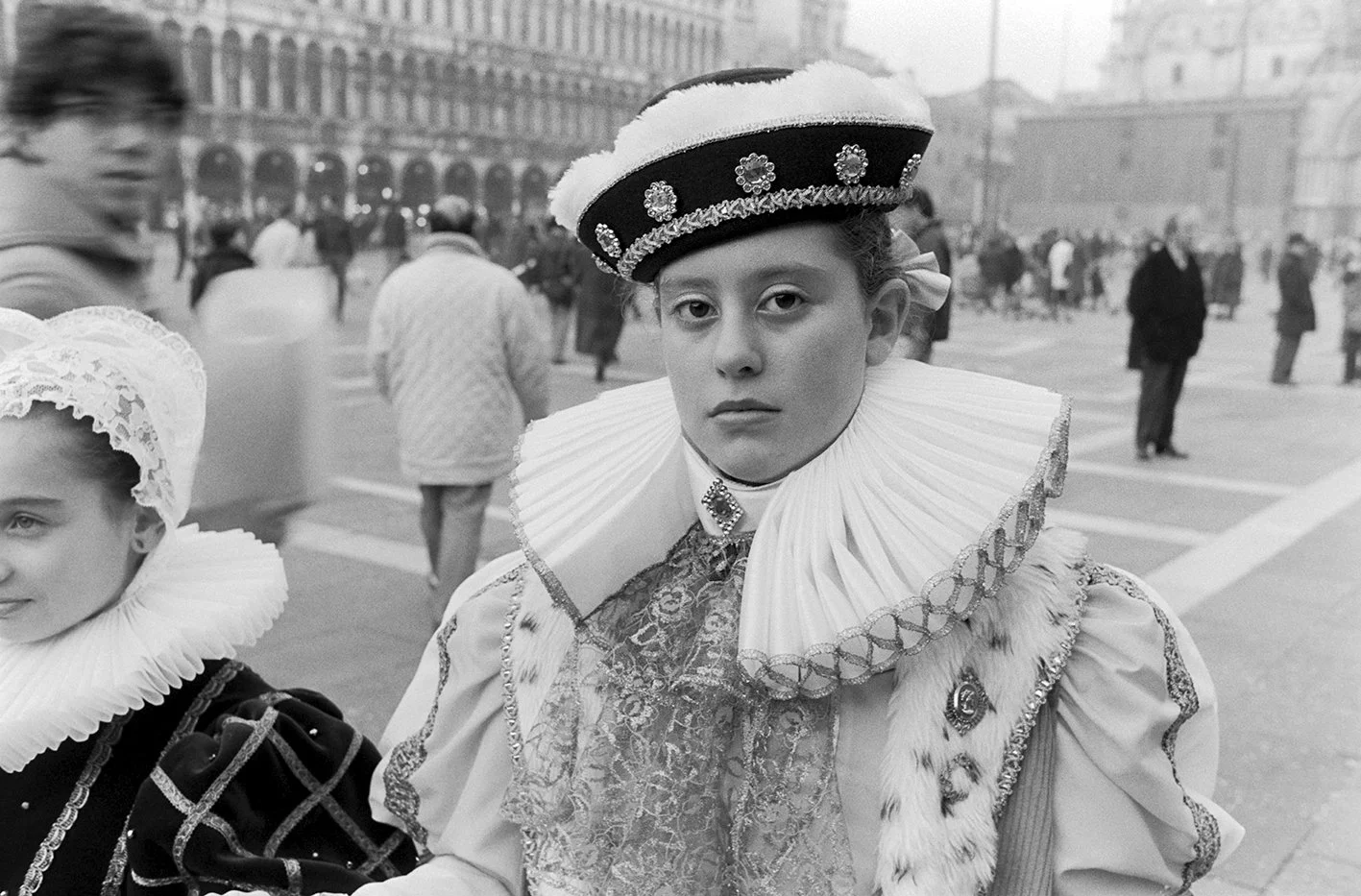 A young person dressed in traditional Venetian Carnival costume, wearing ornate clothing and a decorative hat, in a busy public square.