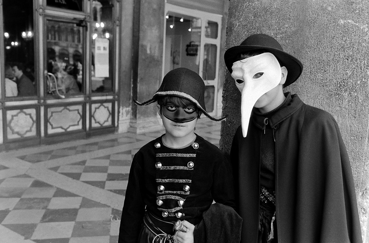 Two children dressed in costumes, one as a soldier with a helmet and mask, and the other as a plague doctor with a bird-like mask and a cape, standing indoors against a wall.