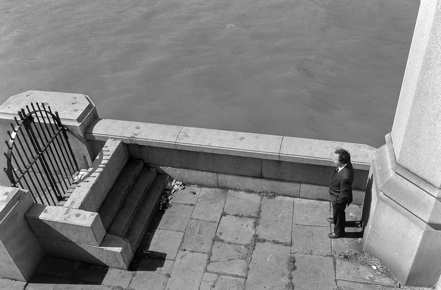 A man in a suit standing on a stone promenade near a river, with stairs leading down to the water and a wrought iron gate at the top of the stairs.