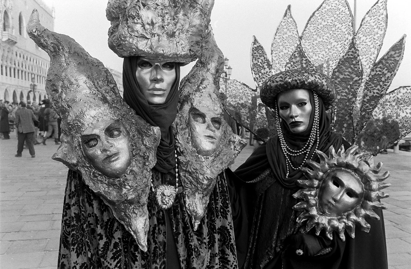 Two people in elaborate costumes with masks and headpieces, standing outdoors in a city square, possibly during a festival or parade. The costumes are ornate, themed with faces and natural elements.