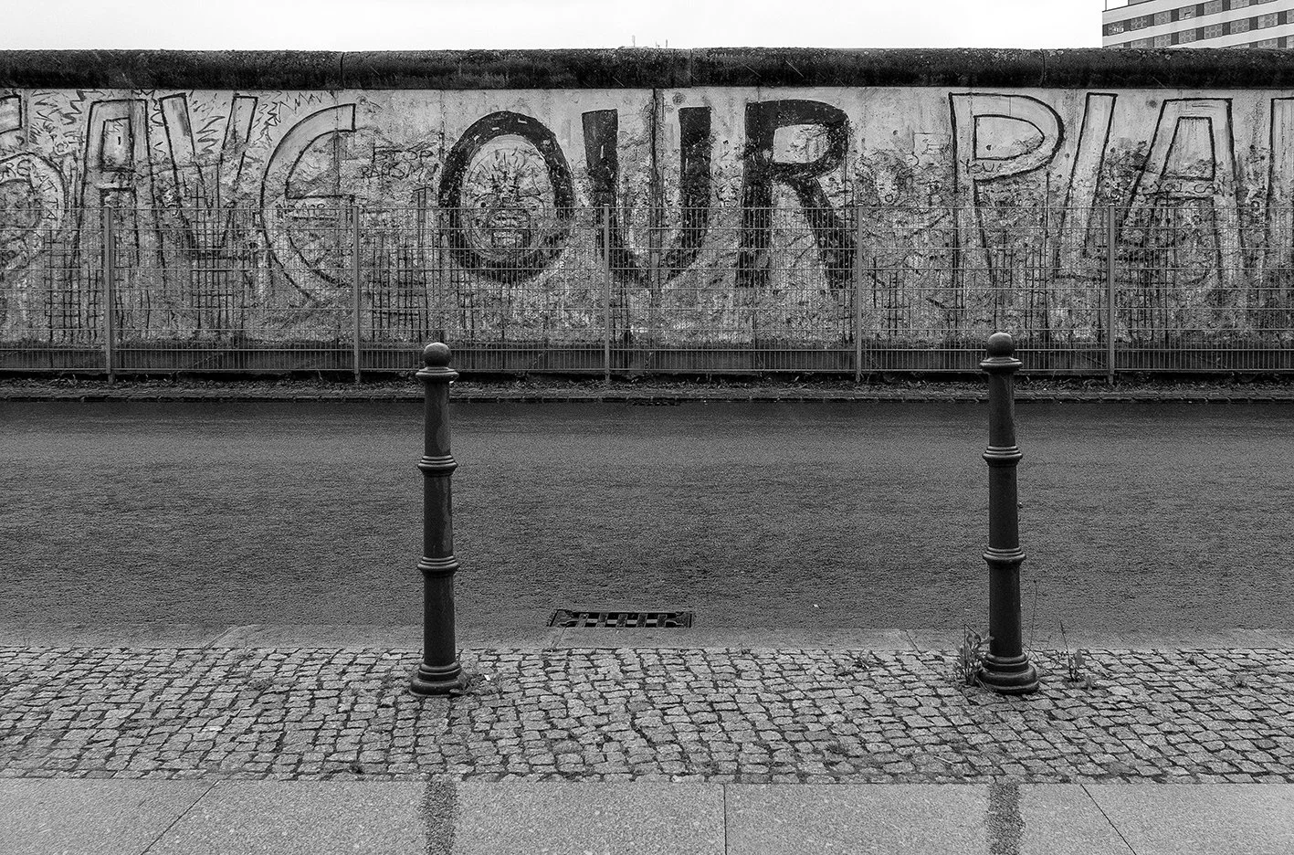Black and white photograph of a graffiti-covered concrete wall with the word 'CONCRETE' painted on it. In the foreground, there are two metal bollards on a cobblestone sidewalk, with a small drain in the pavement, and no people are visible.