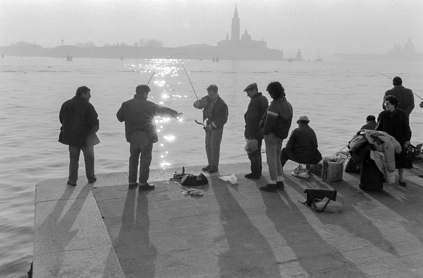 People fishing on a concrete pier by the water with a distant cityscape and church spire in the background.