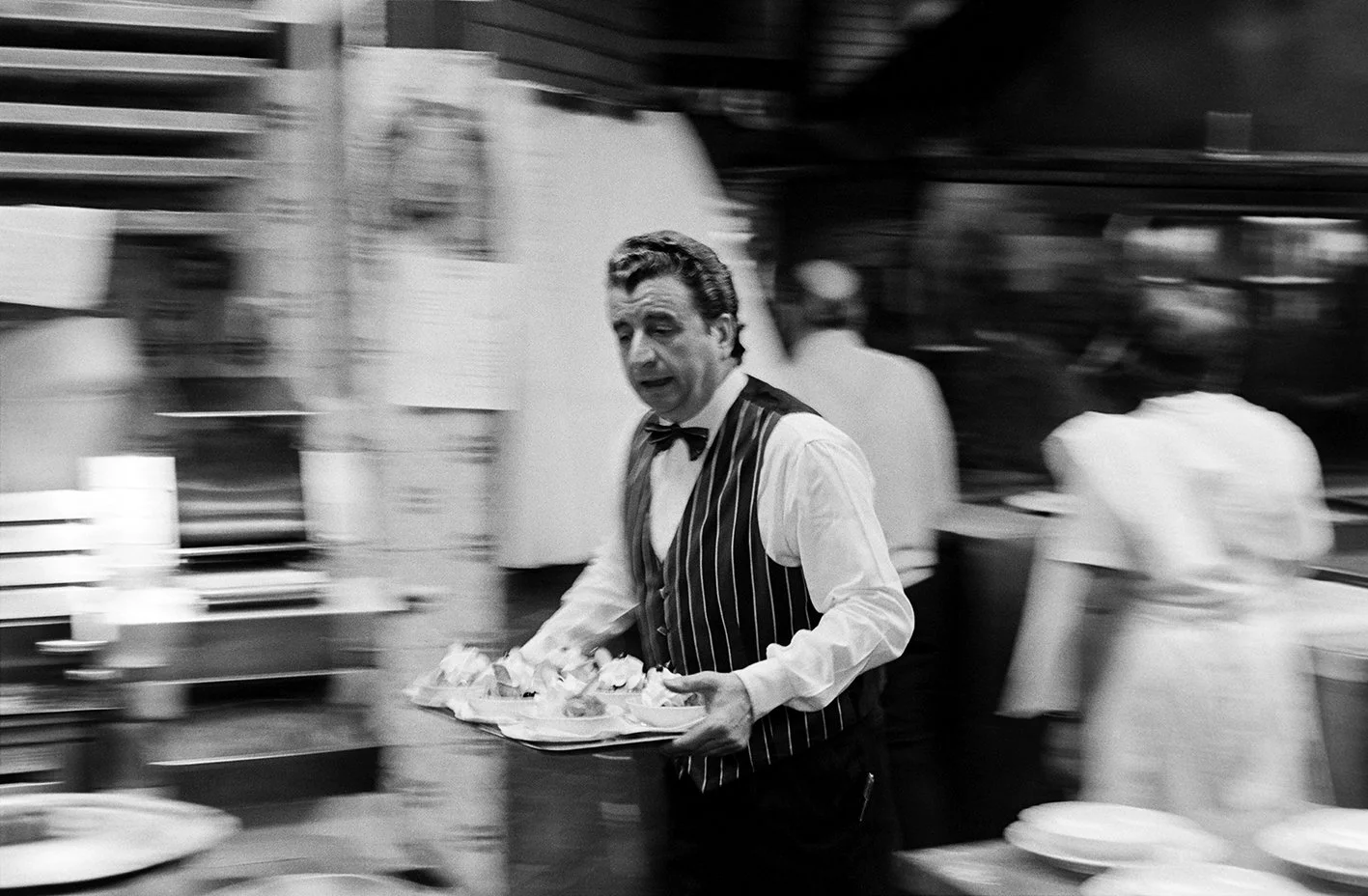 A waiter in a black and white pinstripe vest and bow tie carries a tray with plates of food in a busy restaurant kitchen.