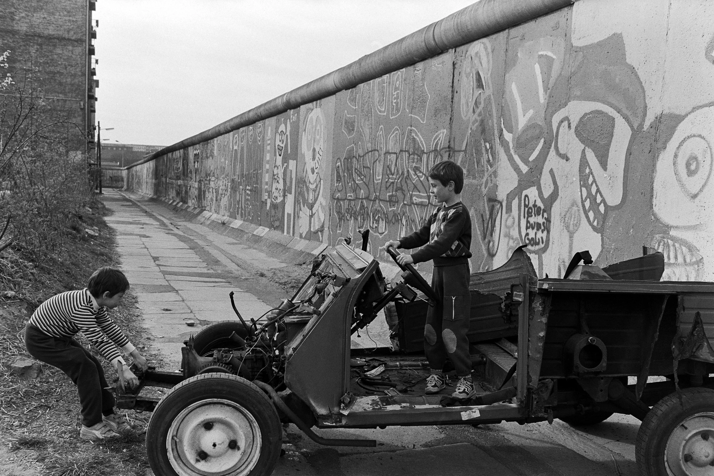 Two boys working on a demolition project near a graffitied wall, one boy is under the truck working on the wheel while the other is standing on the truck's platform. The scene is outdoors along a sidewalk next to a graffiti-covered wall.