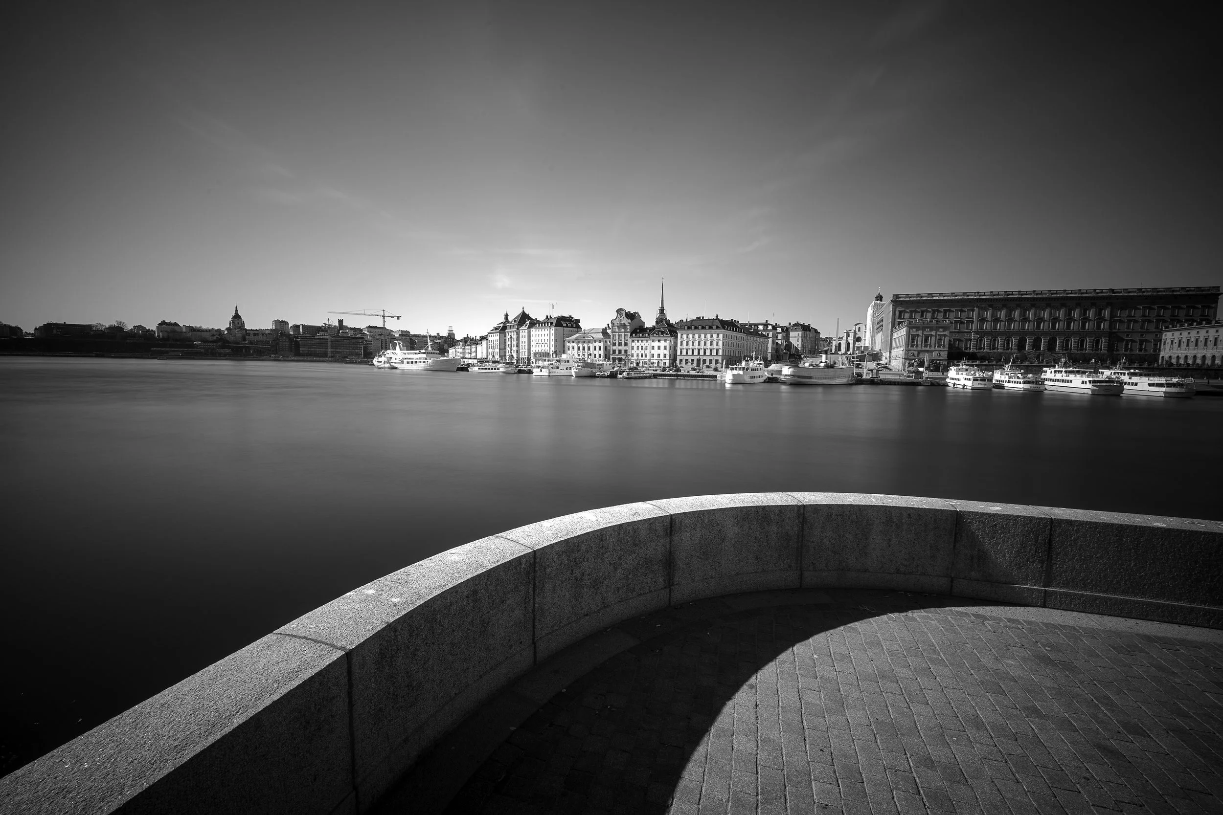 Black and white photo of a city skyline across a river, with buildings and boats docked along the waterfront, viewed from a curved stone promenade.