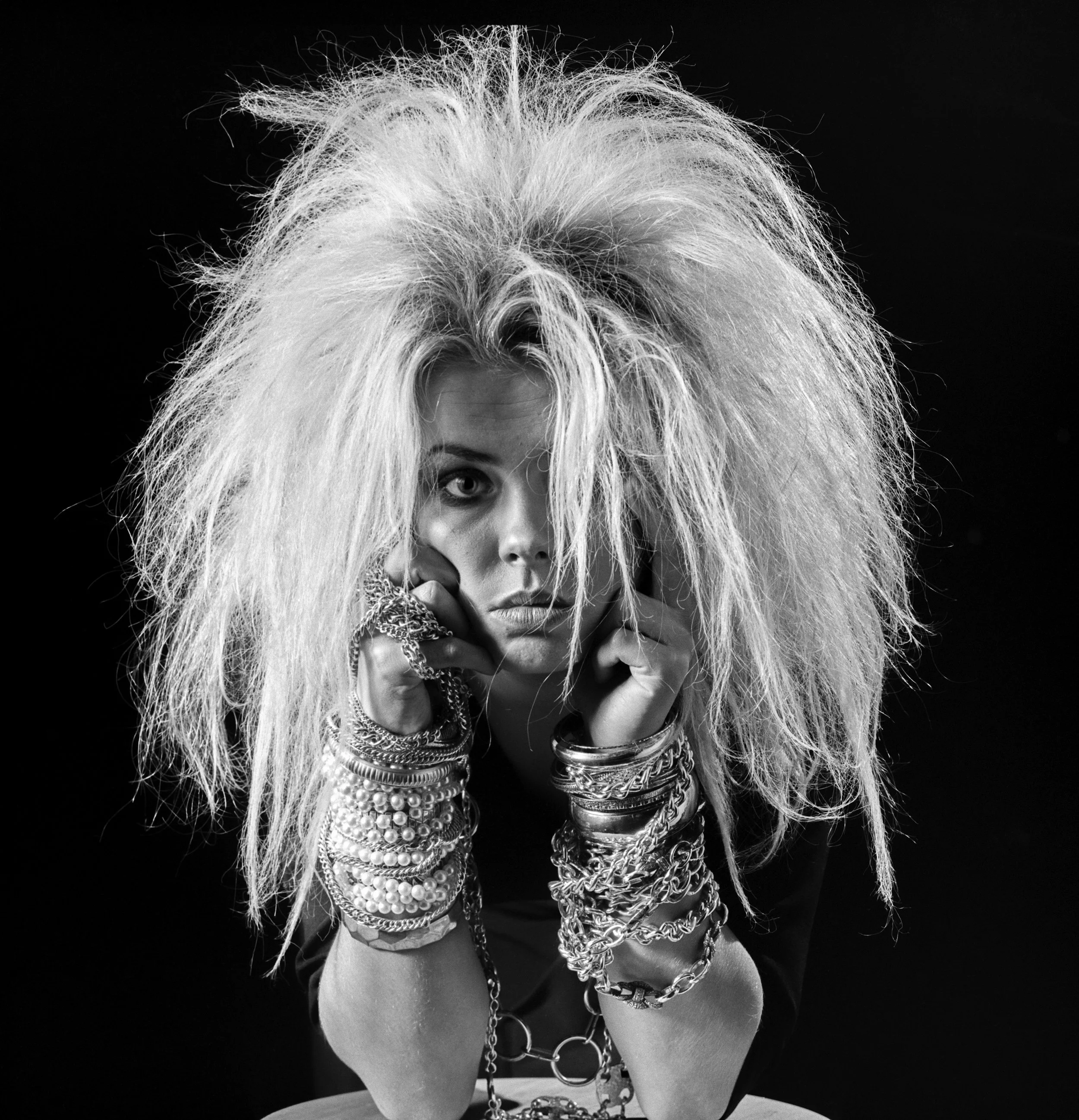 Black and white photo of woman with wild, frizzy blonde hair, wearing multiple jewelry pieces and resting her head on her hands.
