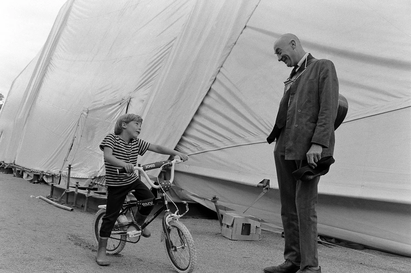 A young girl on a bicycle interacting with an older man standing nearby, in front of a large tent.
