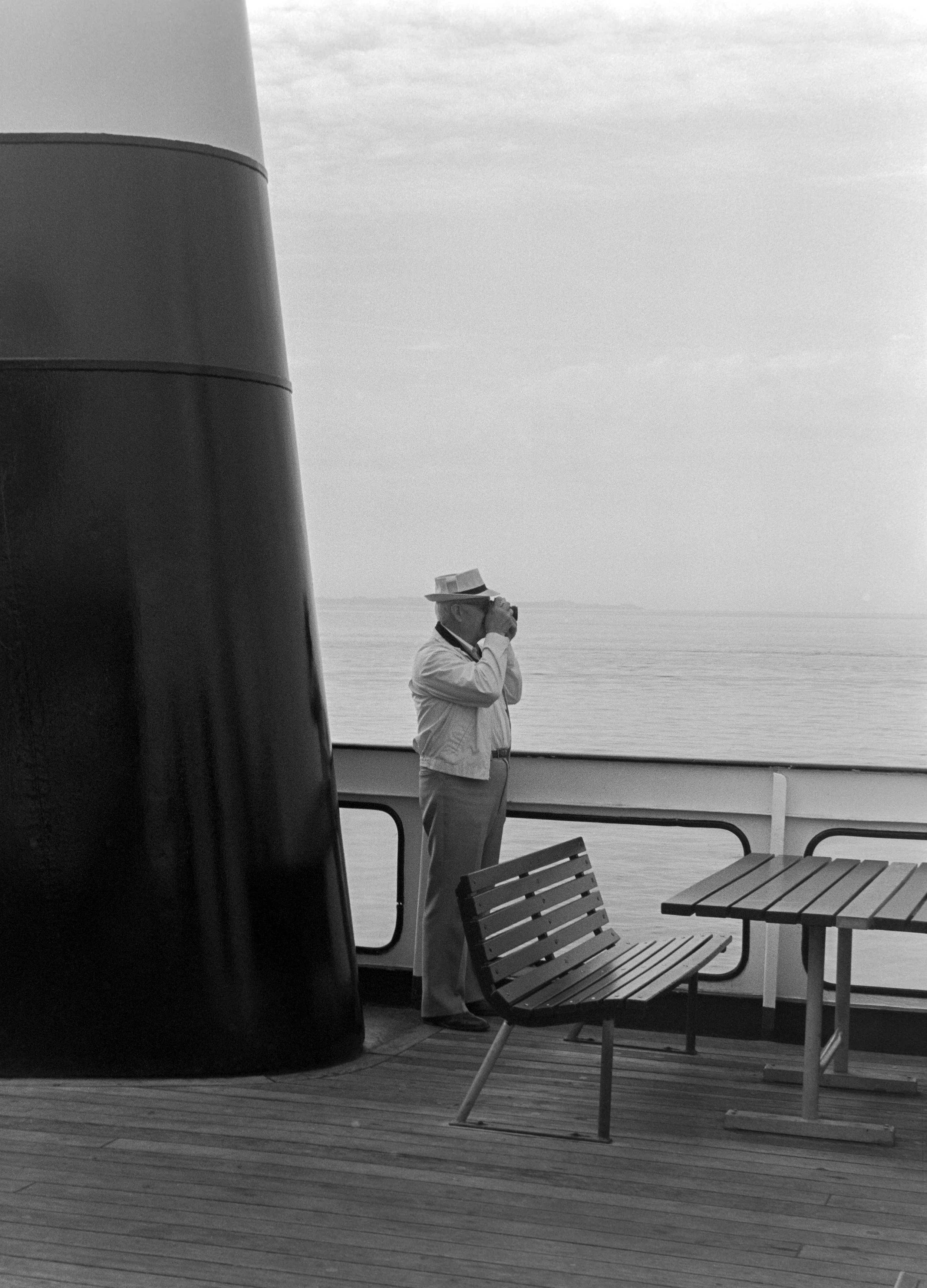 A man wearing a hat and a jacket stands on the deck of a ship, taking a photograph with a camera. The deck has wooden flooring, benches, and a railing overlooking a calm body of water, with a cloudy sky above.