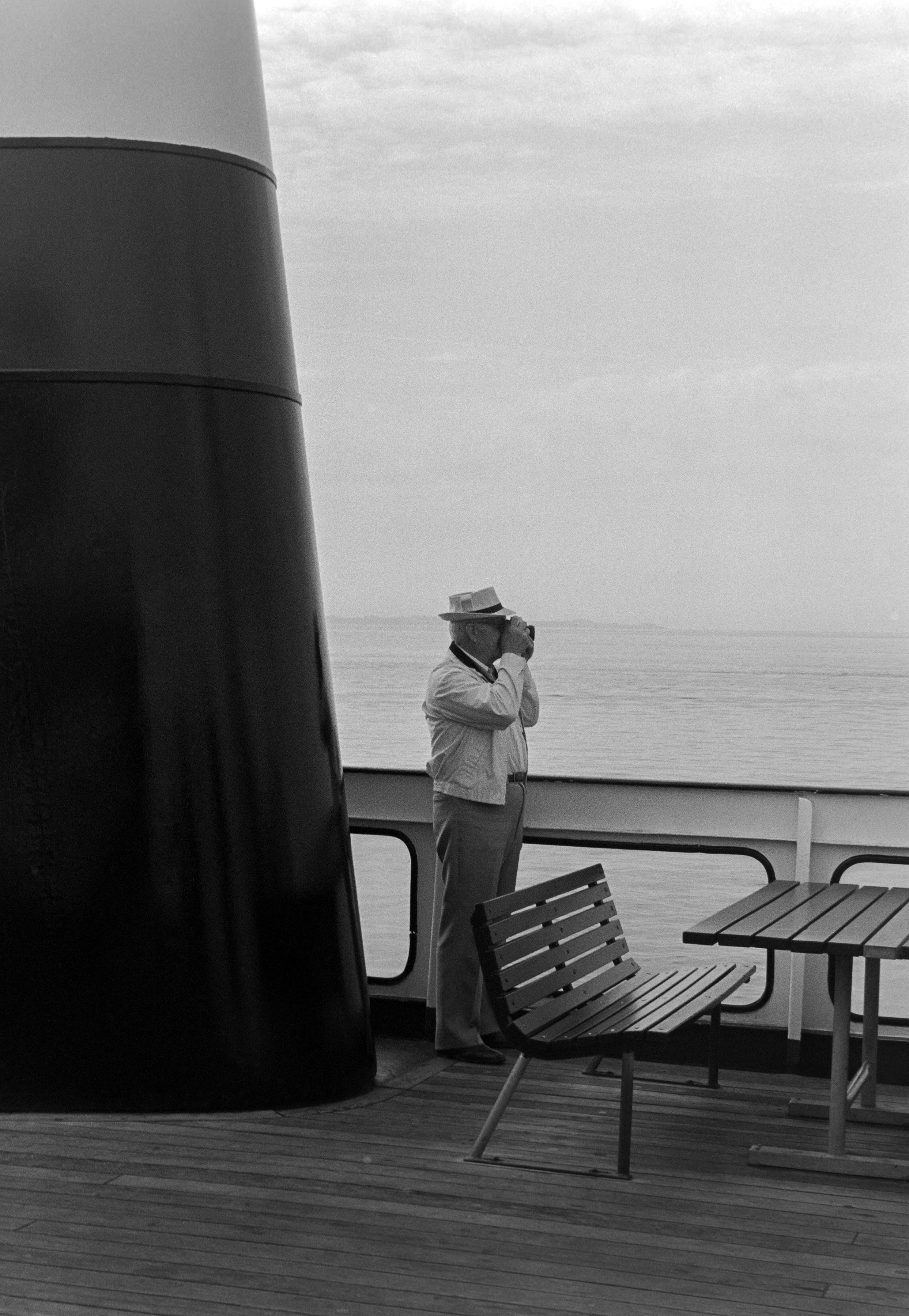A man wearing a hat and jacket, standing on a boat deck, taking a photo with a camera, with the sea and sky in the background.