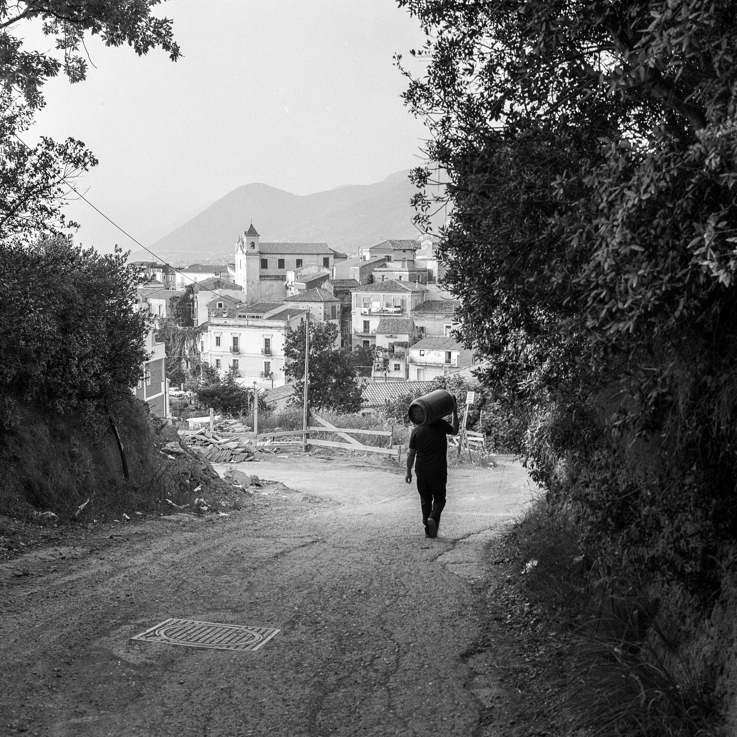 A person walking down a dirt road carrying a large bucket on their shoulder, with a town and mountains in the background, in black and white.