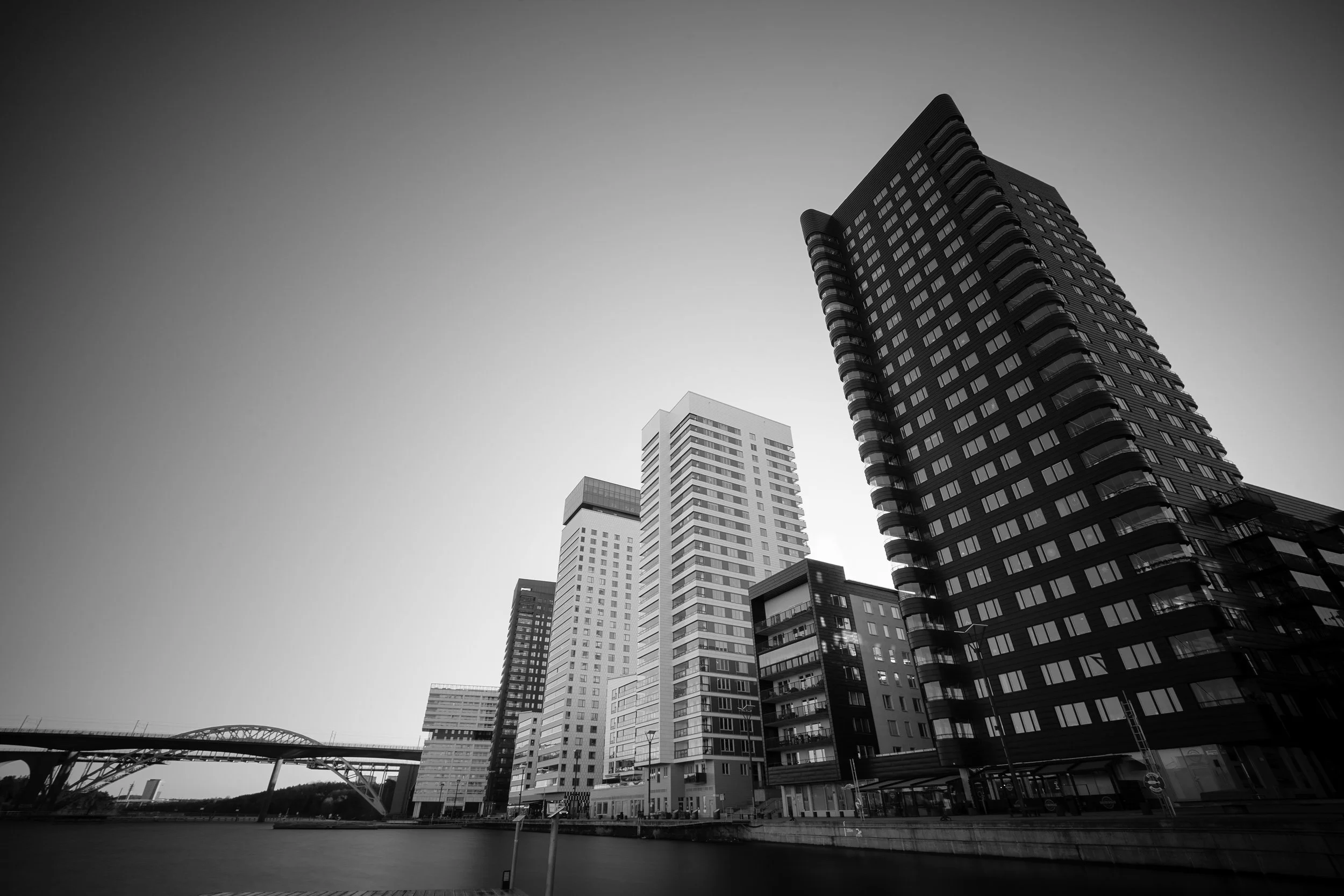 Black and white photo of modern high-rise buildings along a waterway with a bridge in the background.