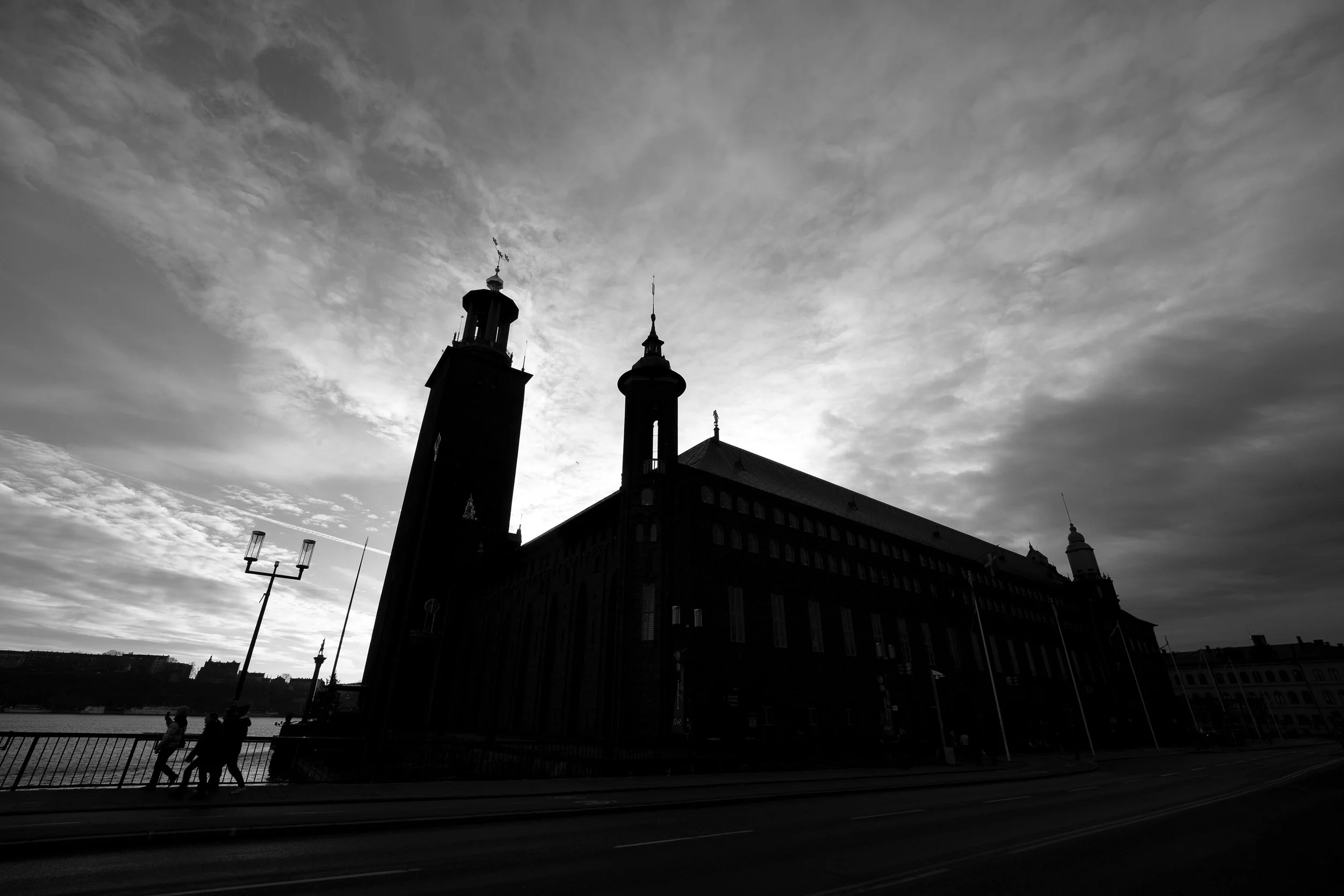 Silhouette of a historic church or cathedral with tall towers against a cloudy sky, near a waterfront with people walking by.