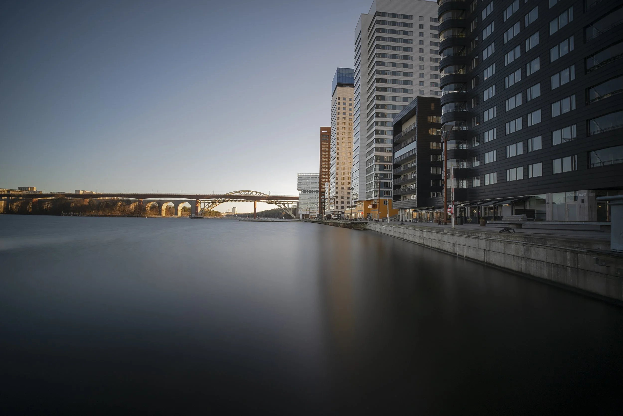 Cityscape with high-rise buildings along a river, with a bridge in the distance during sunset or sunrise.