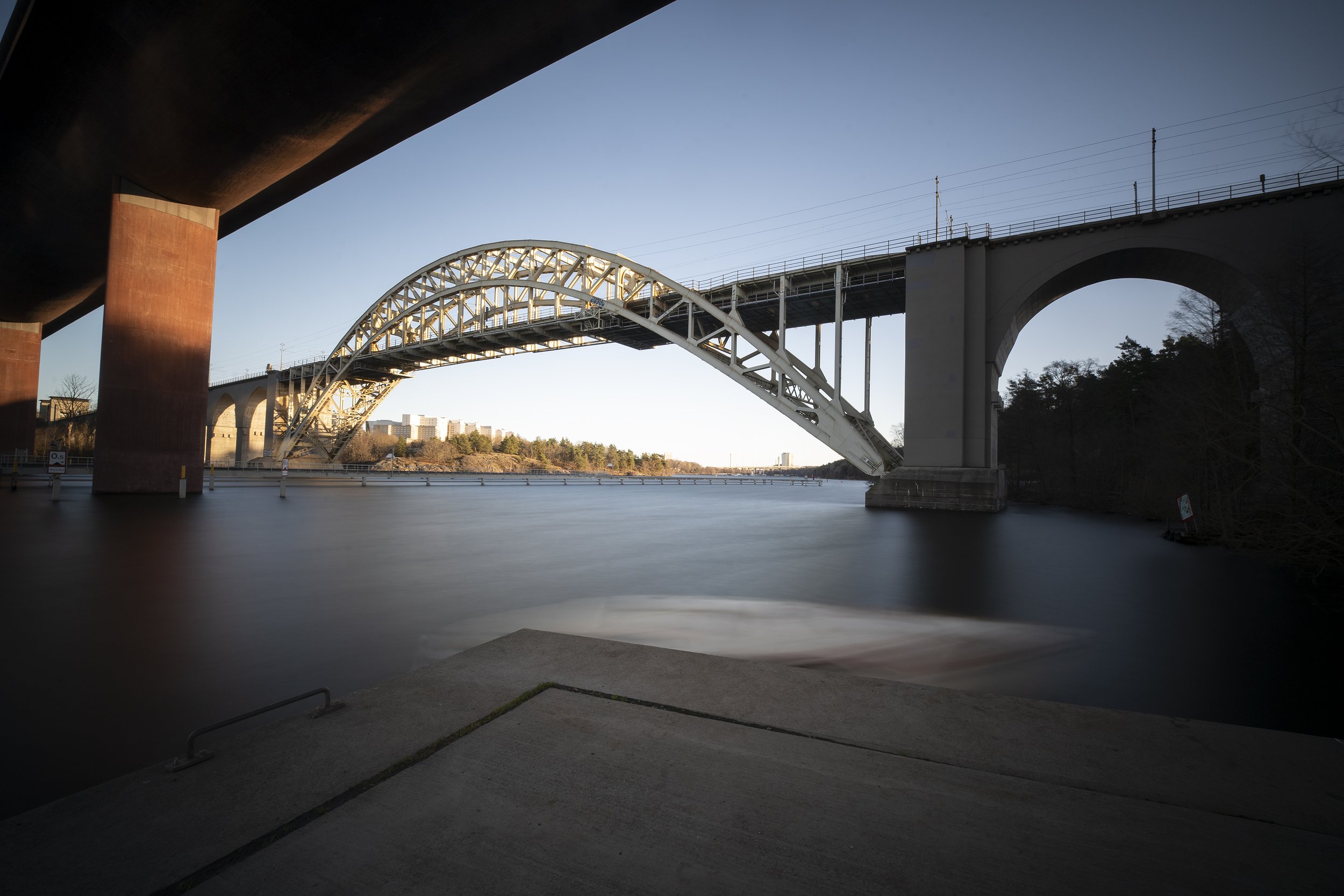 A large metal arch bridge spanning over a river, with the photo taken from beneath a nearby structure during daylight hours.