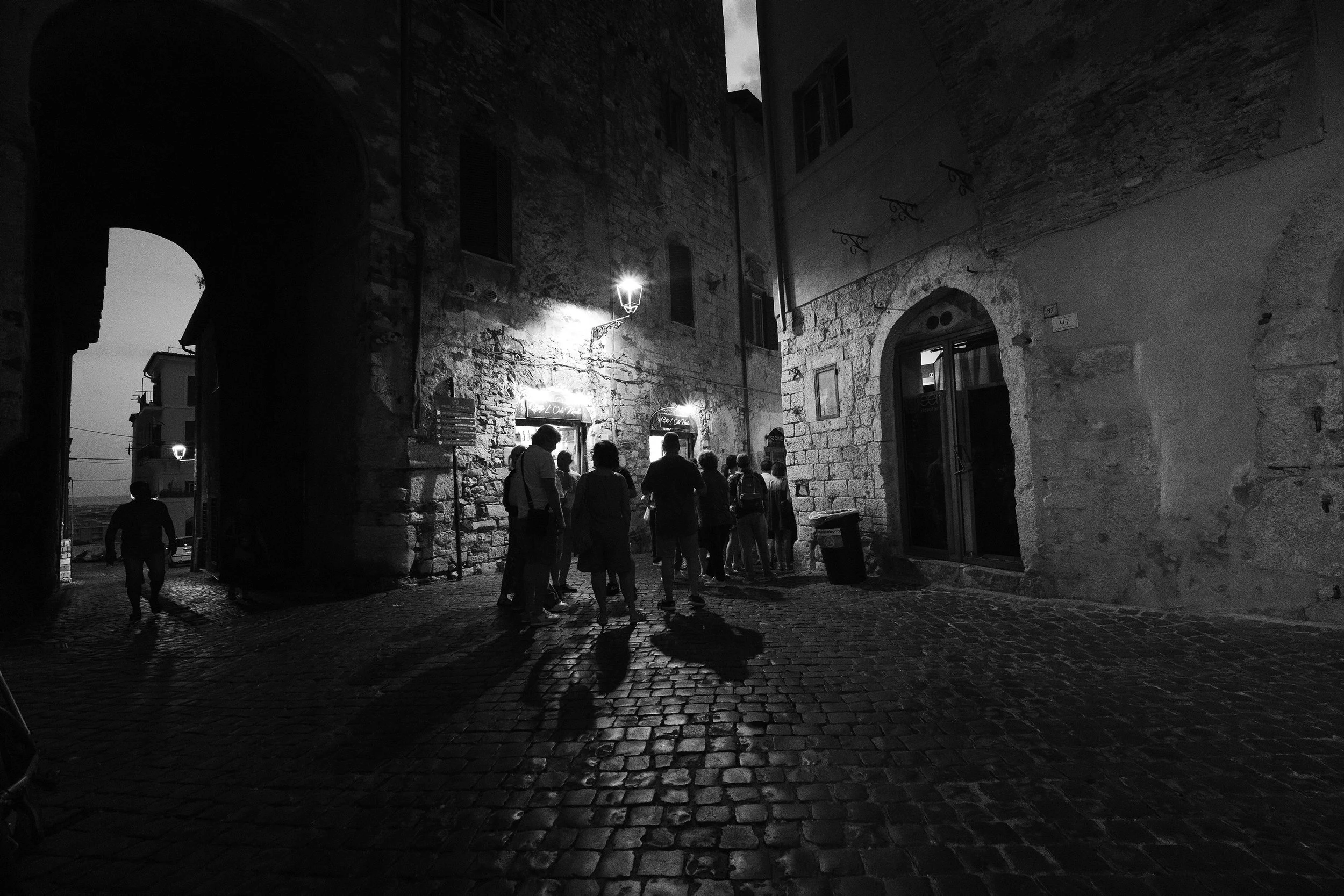 A nighttime scene of a narrow cobblestone street with groups of people gathered near a building with illuminated signs, surrounded by old stone buildings. A street lamp lights up part of the street.