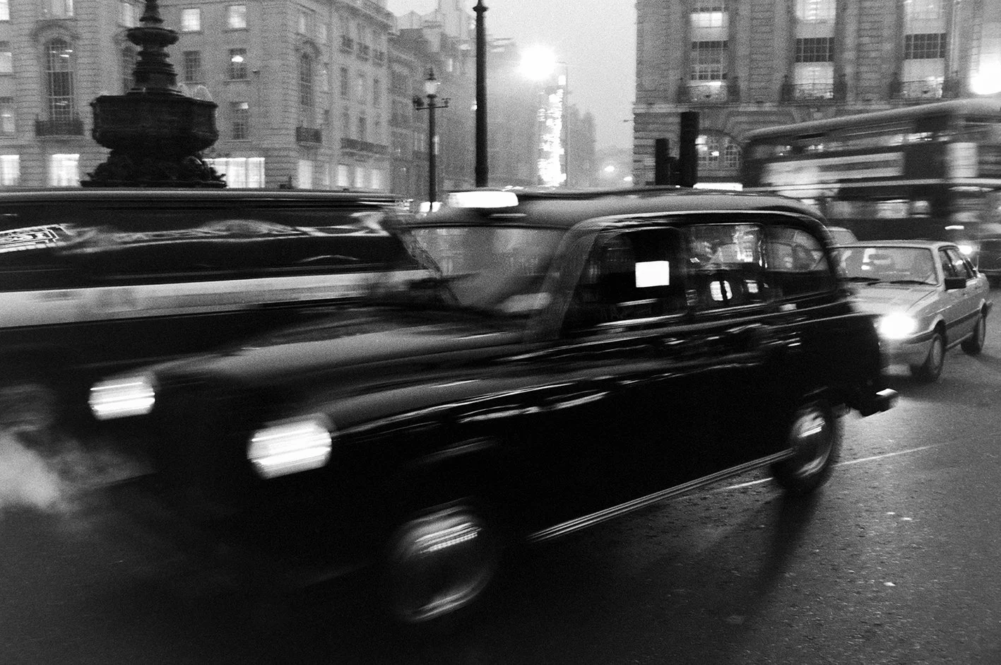 Black and white photo of cars driving on a city street at night, with buildings and streetlights in the background.