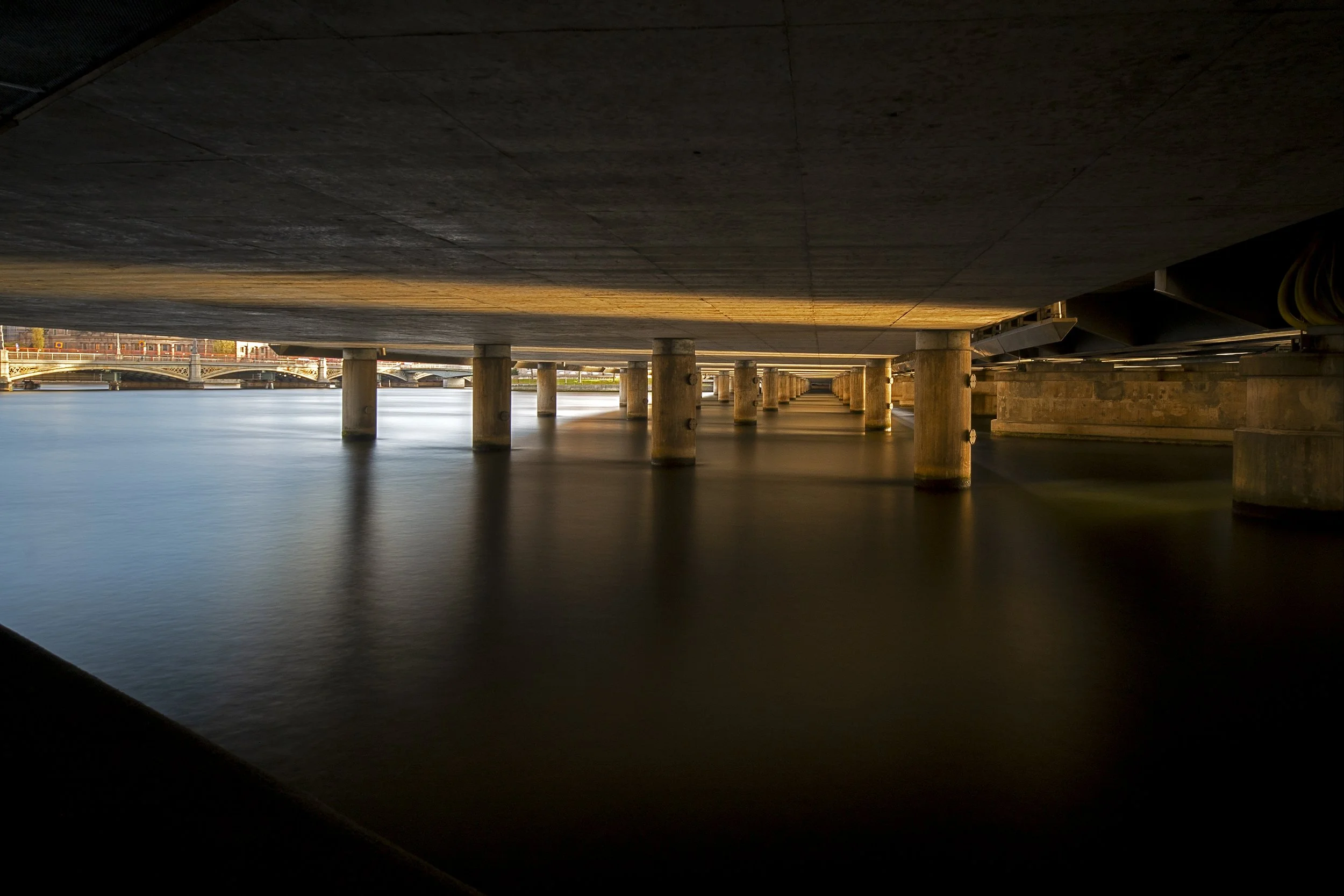 Underneath a bridge over a river, showing the bridge's understructure with support columns. The water is calm and reflects the elements above. In the background, part of a cityscape with buildings and a bridge skyline is visible.