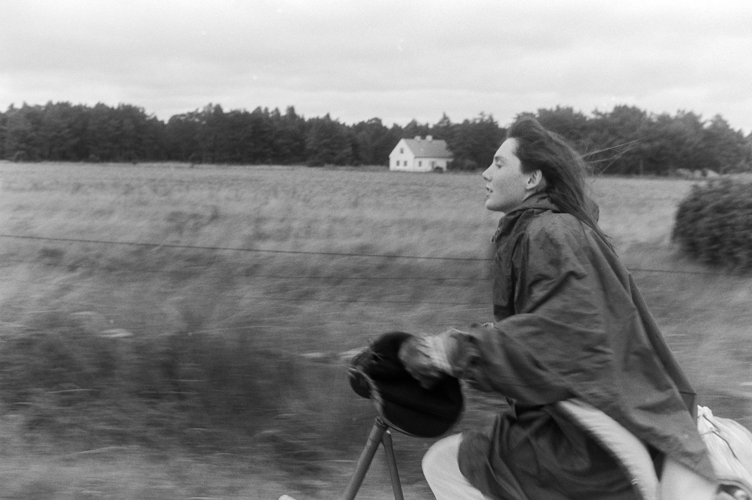 A woman with long hair flowing in the wind, riding a bicycle through a rural landscape with open fields and a house in the distance, in black and white.