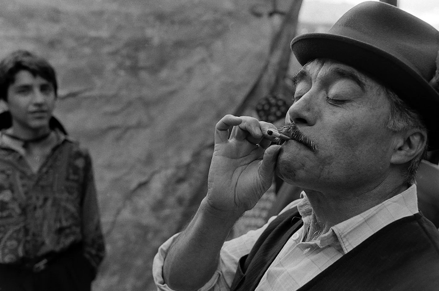 A man wearing a hat with closed eyes, smoking a cigarette, and a young man with short hair in the background, standing outdoors.