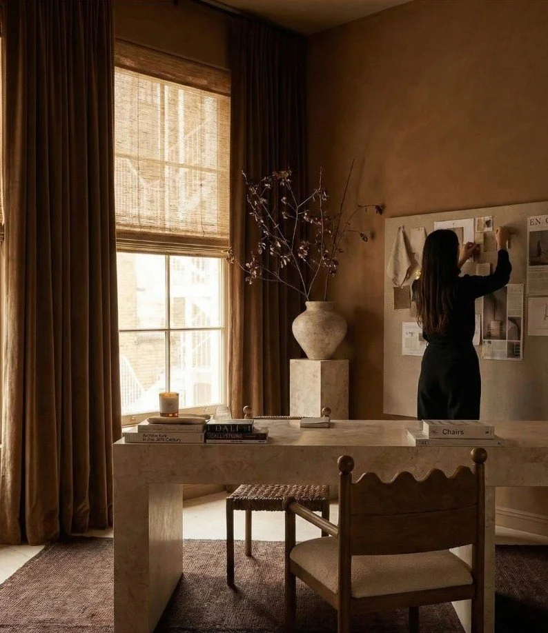 Woman in black dress pinning papers or photos on a corkboard in a sunlit room with beige walls, a large window with curtains, a marble table, a chair, a large vase with branches, and books and candles on the table.