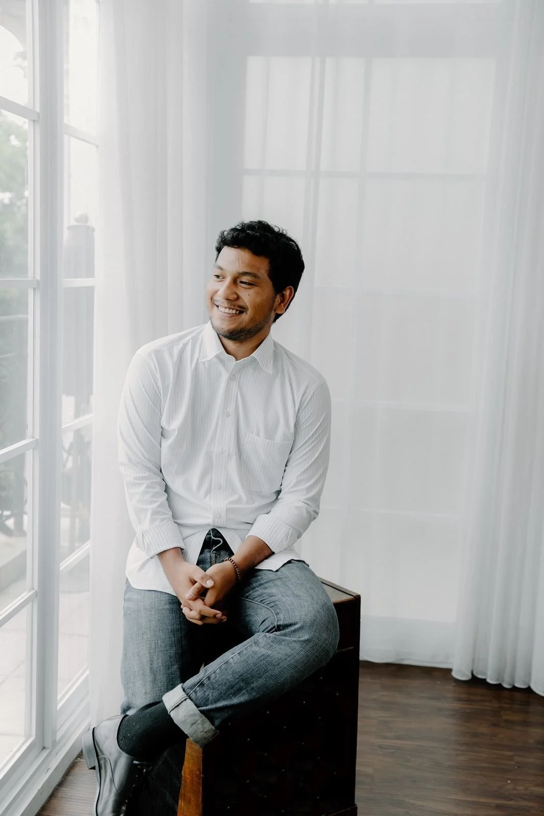 A man sitting on a wooden stool near a large window with white curtains, smiling and looking to his left, wearing a white button-up shirt, gray jeans, and black shoes.