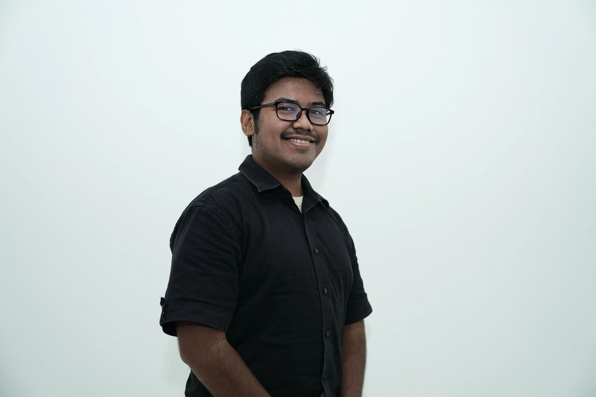 Smiling young man with dark hair and glasses, wearing a black shirt, standing against a plain white background.