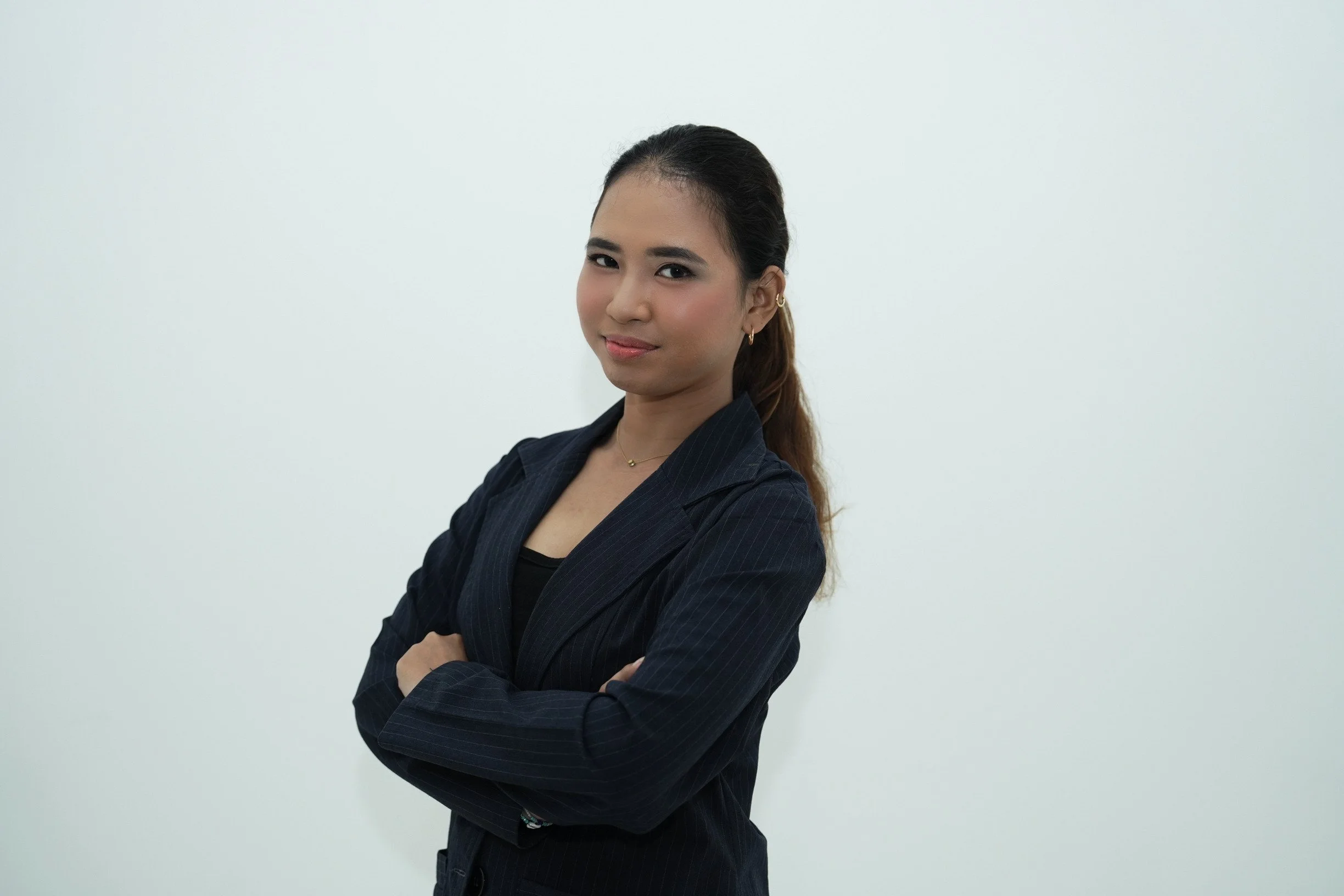 A young woman with dark hair tied back, wearing a dark pinstripe blazer over a black top, standing with arms crossed against a plain white wall.