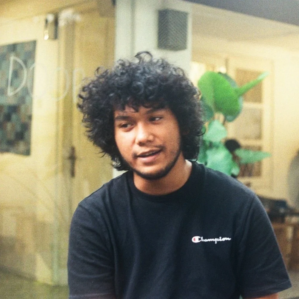 A young man with curly hair wearing a black Champion t-shirt, sitting indoors with a green plant in the background.