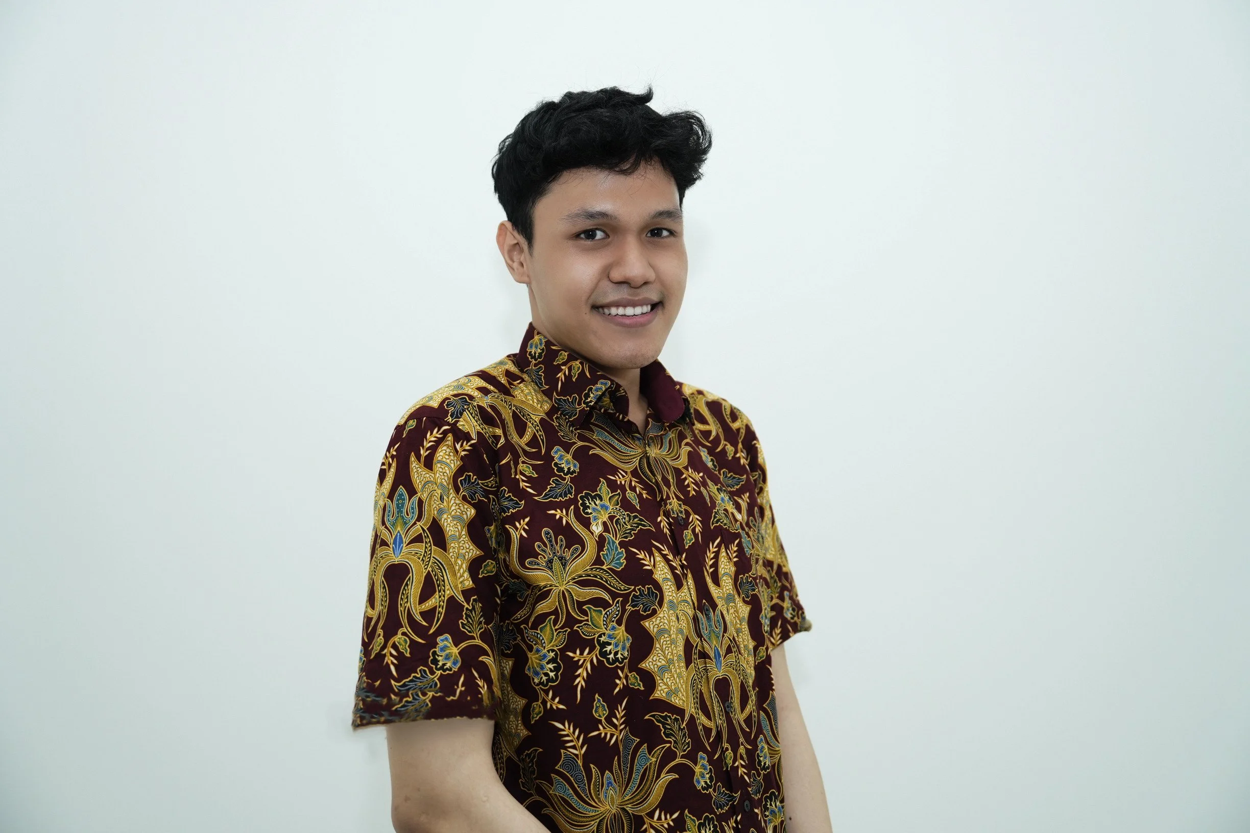A young man smiling and wearing a traditional dark-colored batik shirt with gold, blue, and red patterns, standing in front of a plain white background.