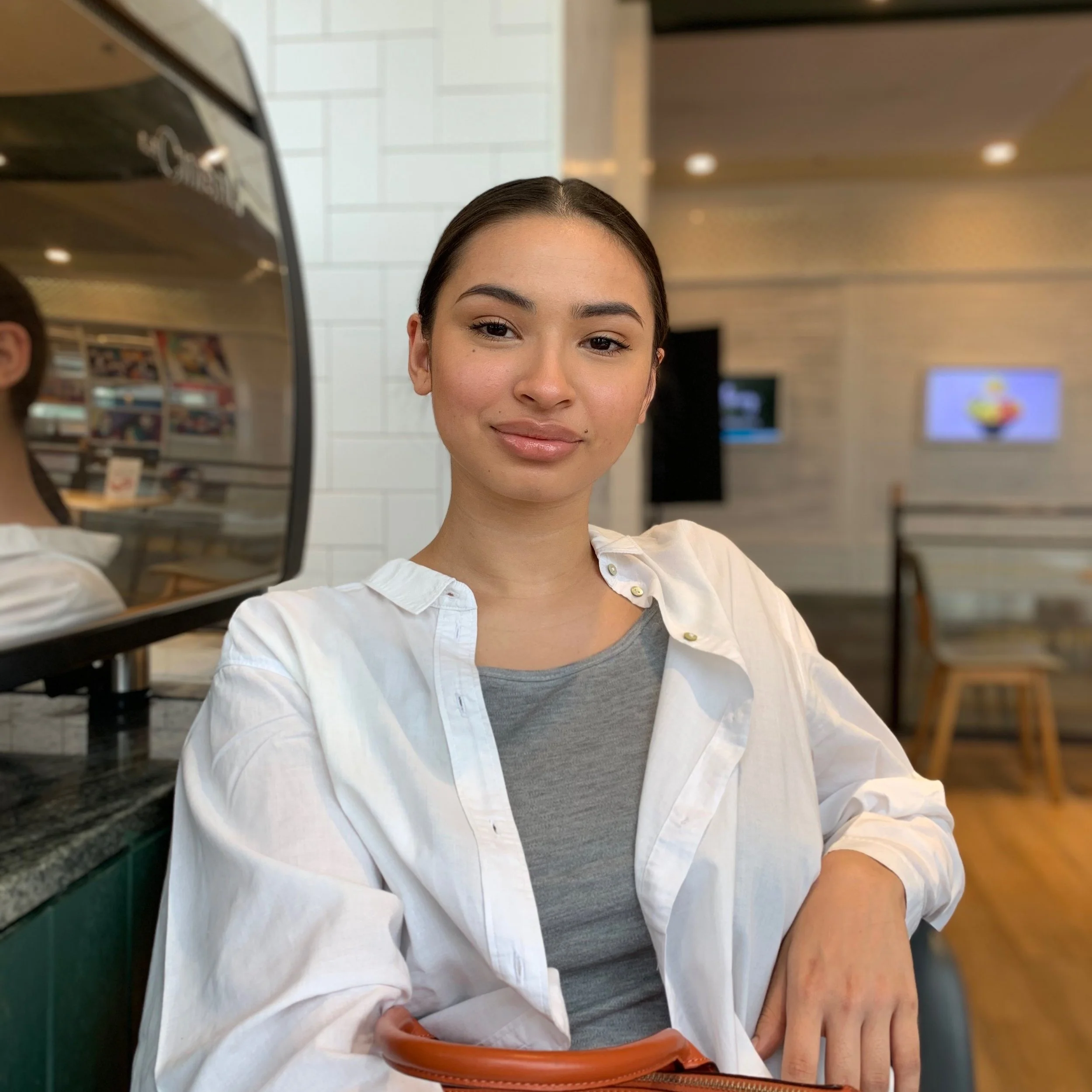Young woman with dark hair, wearing a white shirt over a gray top, sitting in a cafe or restaurant with a reflective surface beside her and digital screens in the background.
